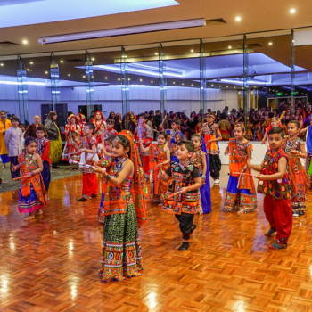 Children in colourful traditional Indian outfits dancing with sticks at an indoor event.